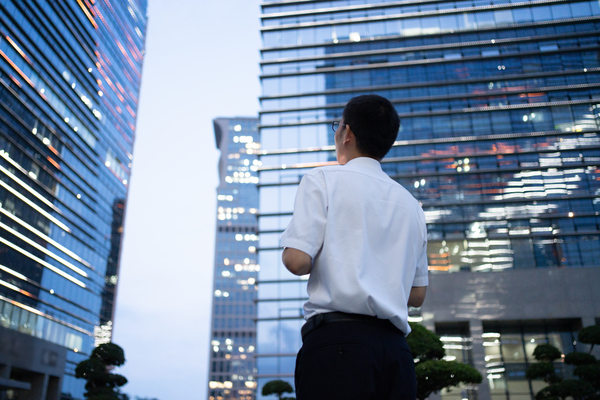 A young Asian business man in a white shirt looks up at tall buildings